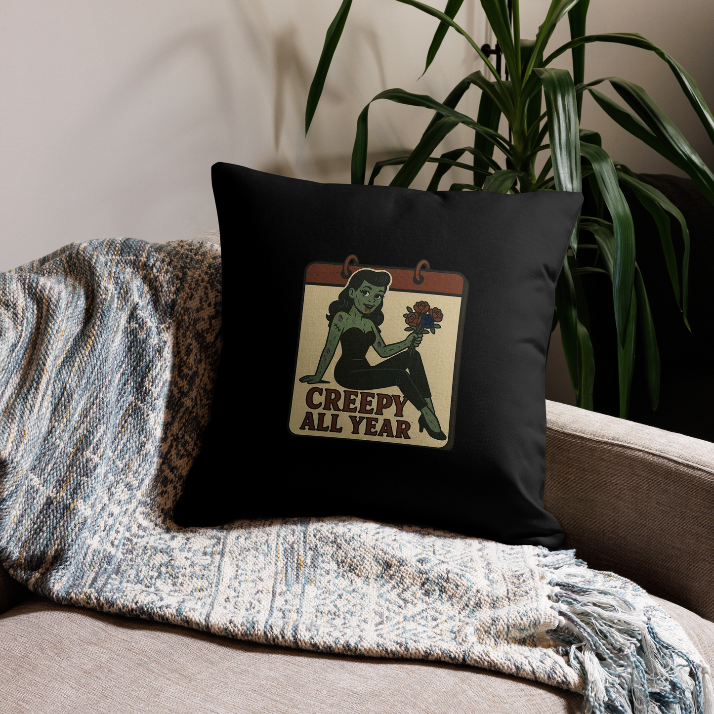 Black pillow with a graphic design on a couch next to a plant. The design features a green pinup-style zombie girl holding flowers, with a banner underneath that reads “CREEPY ALL YEAR.”