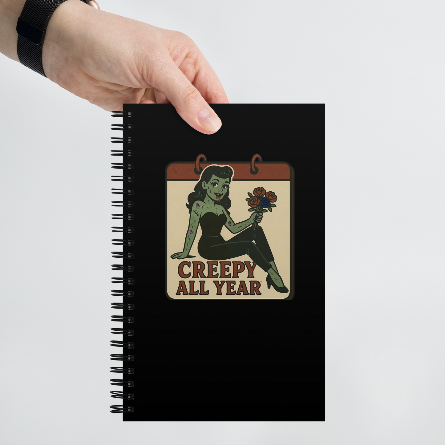Black notebook with a pinup zombie holding flowers design and 'Creepy All Year' text, held by a hand on a light gray background.