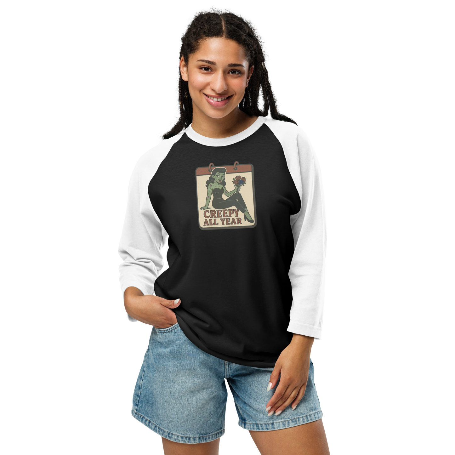 A woman with black hair wearing jean shorts and black and white raglan shirt. The design features a green pinup-style zombie girl holding flowers, with a banner underneath that reads “CREEPY ALL YEAR.”