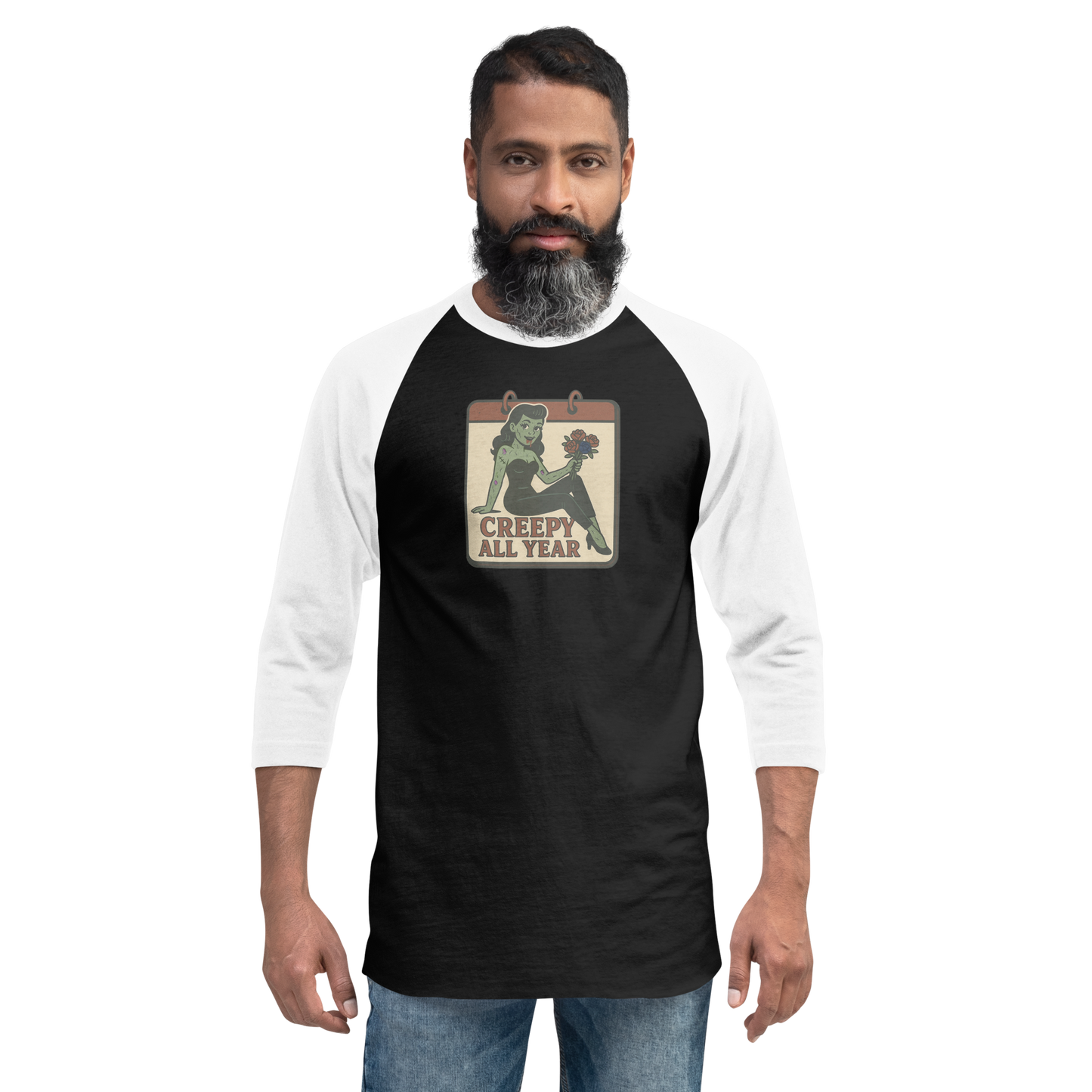 A man with black hair wearing jeans and a black and white raglan t-shirt with a small vintage-style design on the pocket area. The design features a green pinup-style zombie girl holding flowers, with a banner underneath that reads “CREEPY ALL YEAR.”