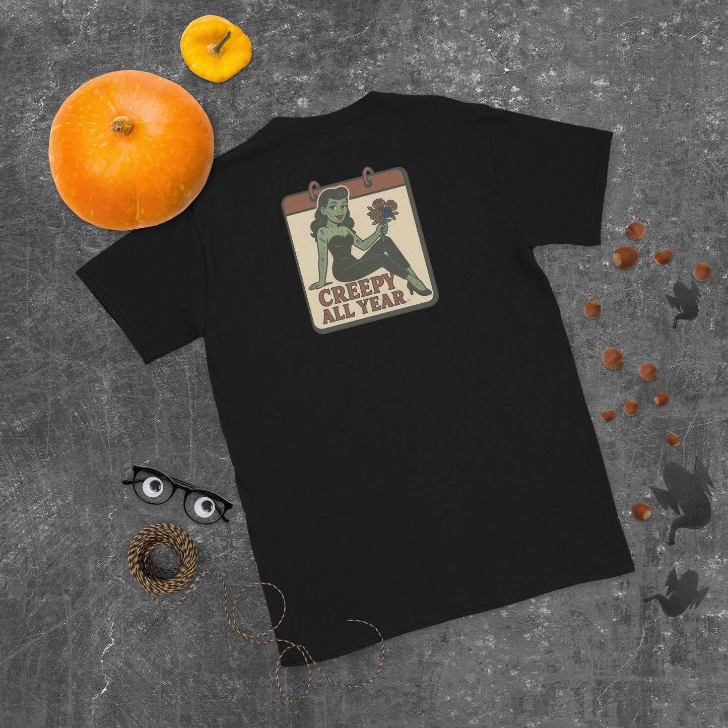 A black t-shirt with a vintage-style design on the back area surrounded by a dark grey pattern pumpkins and ghosts. The design features a green pinup-style zombie girl holding flowers, with a banner underneath that reads “CREEPY ALL YEAR.”