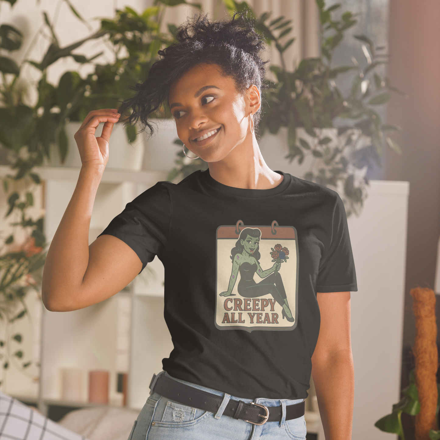 A girl indoors surrounded by plants holding the ends of her black hair wearing jeans and t-shirt with vintage style design. The design on the front features a green pinup-style zombie girl holding flowers, with a banner underneath that reads “CREEPY ALL YEAR.”