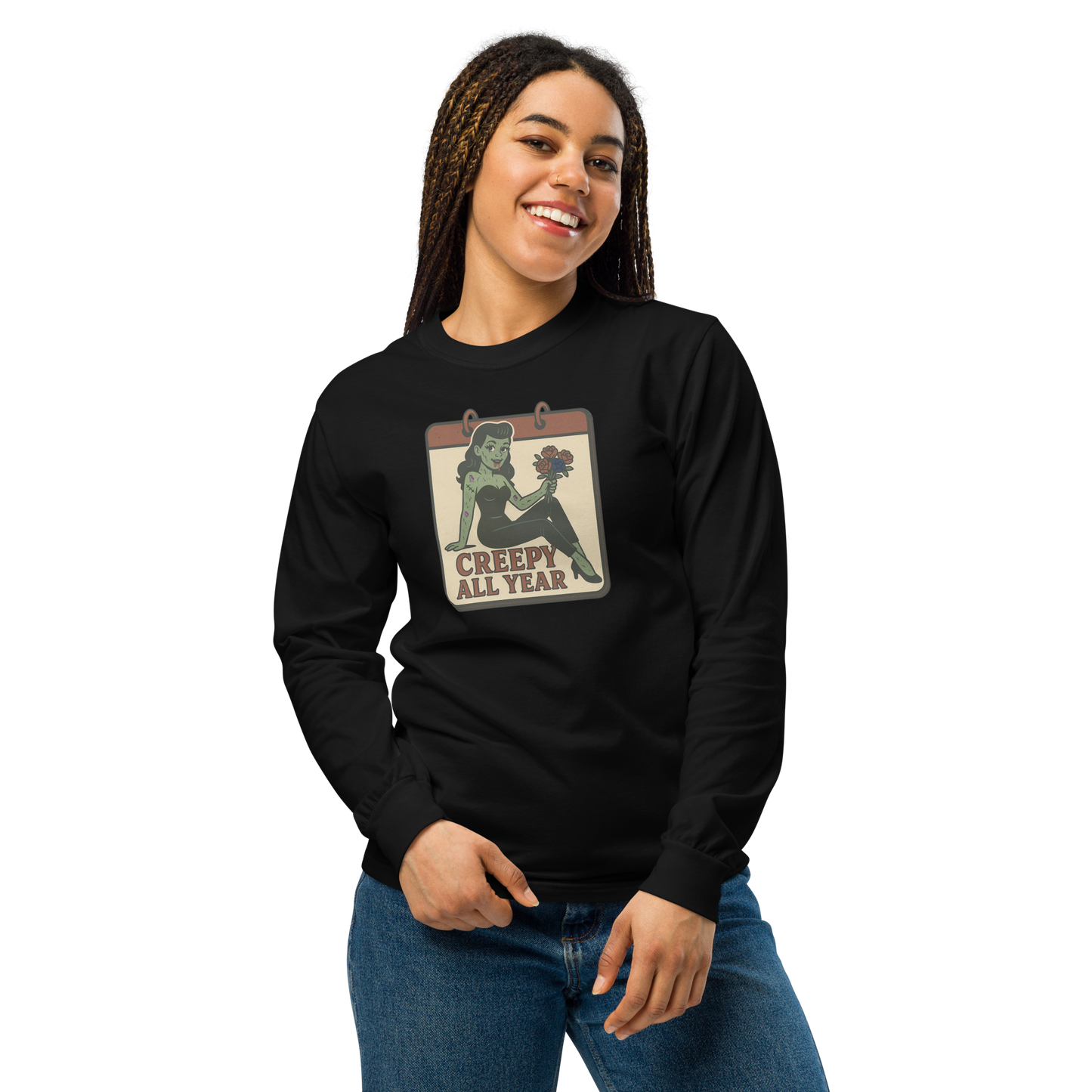 A girl smiling with braids wearing jeans and a black long sleeve t-shirt. The design on the front features a green pinup-style zombie girl holding flowers, with a banner underneath that reads “CREEPY ALL YEAR.”