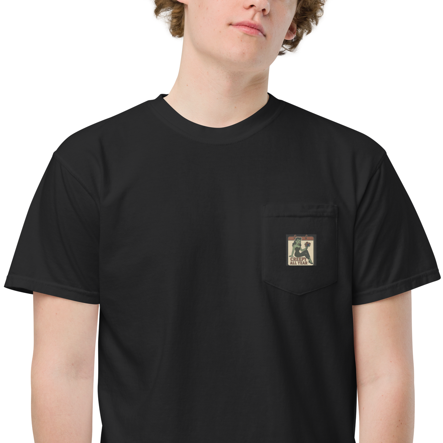 White male with light brown hair wearing a black t-shirt with a small vintage-style patch on the pocket area. The patch features a green pinup-style zombie girl holding flowers, with a banner underneath that reads “CREEPY ALL YEAR.”