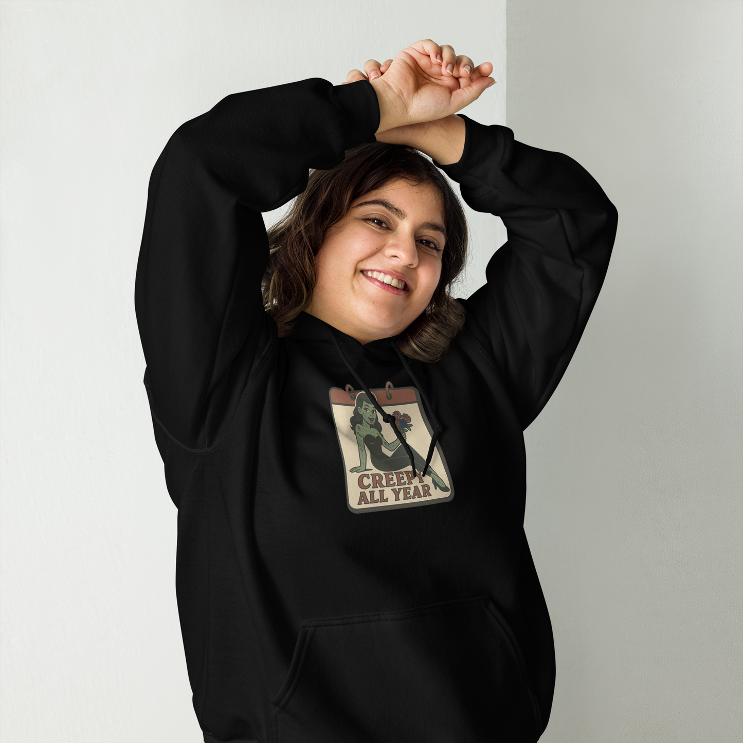 A black hoodie on a girl with brown hair and her hands above her head with a vintage-style design on the front. The design features a green pinup-style zombie girl holding flowers, with a banner underneath that reads “CREEPY ALL YEAR.”