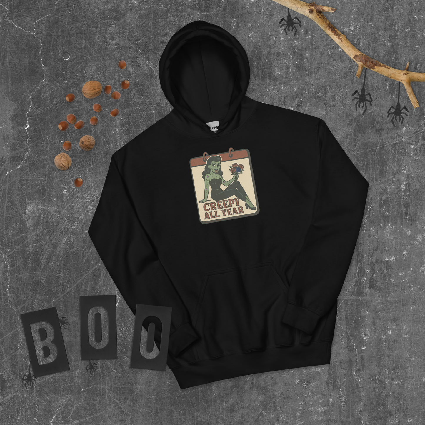 A black hoodie on a grey background surrounded by spiders, with a vintage-style design on the front. The design features a green pinup-style zombie girl holding flowers, with a banner underneath that reads “CREEPY ALL YEAR.”