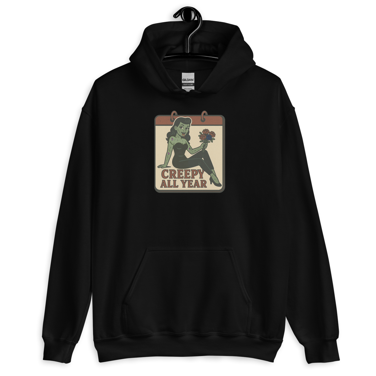 A black hoodie on a hanger with a vintage-style design on the front. The design features a green pinup-style zombie girl holding flowers, with a banner underneath that reads “CREEPY ALL YEAR.”