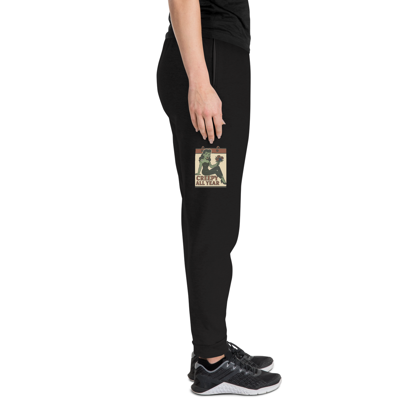 Black joggers on a person with a black t-shirt and grey tennis shoes, with a vintage-style design on the front. The design features a green pinup-style zombie girl holding flowers, with a banner underneath that reads “CREEPY ALL YEAR.”