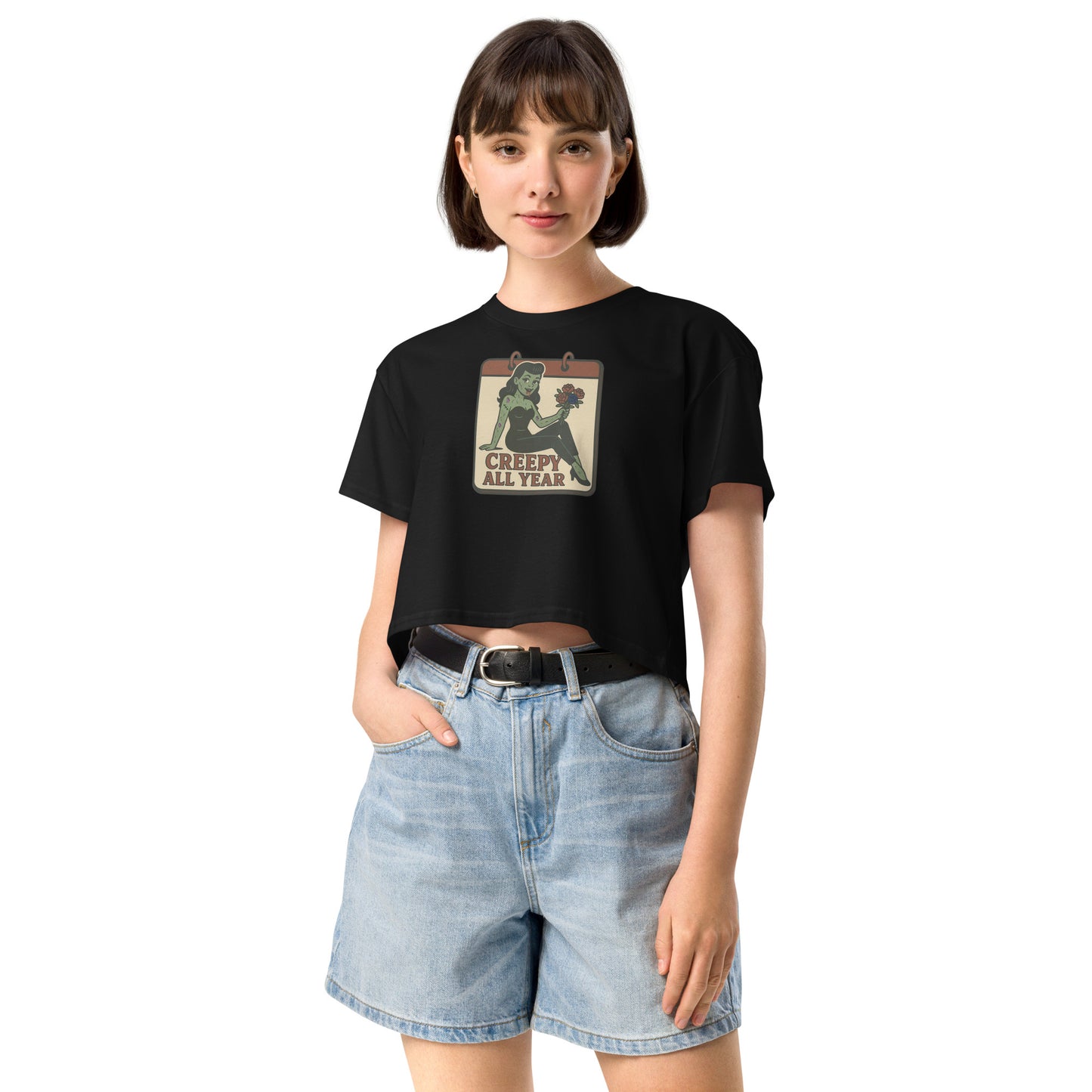 A black crop top on a girl with dark hair and jean shorts with a vintage-style design on the front. The design features a green pinup-style zombie girl holding flowers, with a banner underneath that reads “CREEPY ALL YEAR.”