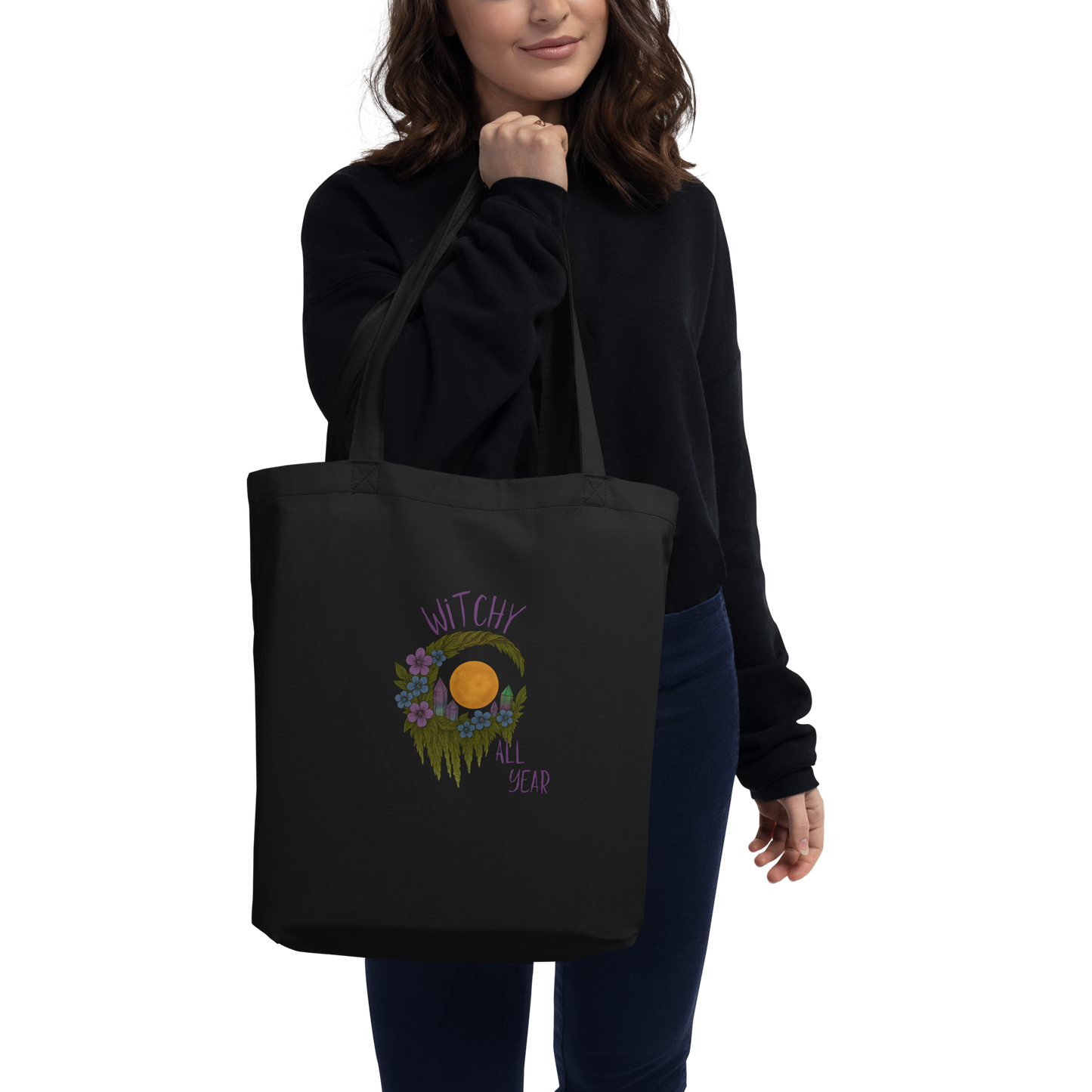 This is a woman with dark hair holding a black eco tote bag with a design of a hand-drawn wreath made of moss-covered branches wraps around a glowing sun. The wreath is decorated with purple and blue flowers, small amethyst and fluorite crystals, and strands of hanging moss. The words “Witchy” curve above the wreath and “All Year” sits on the bottom right