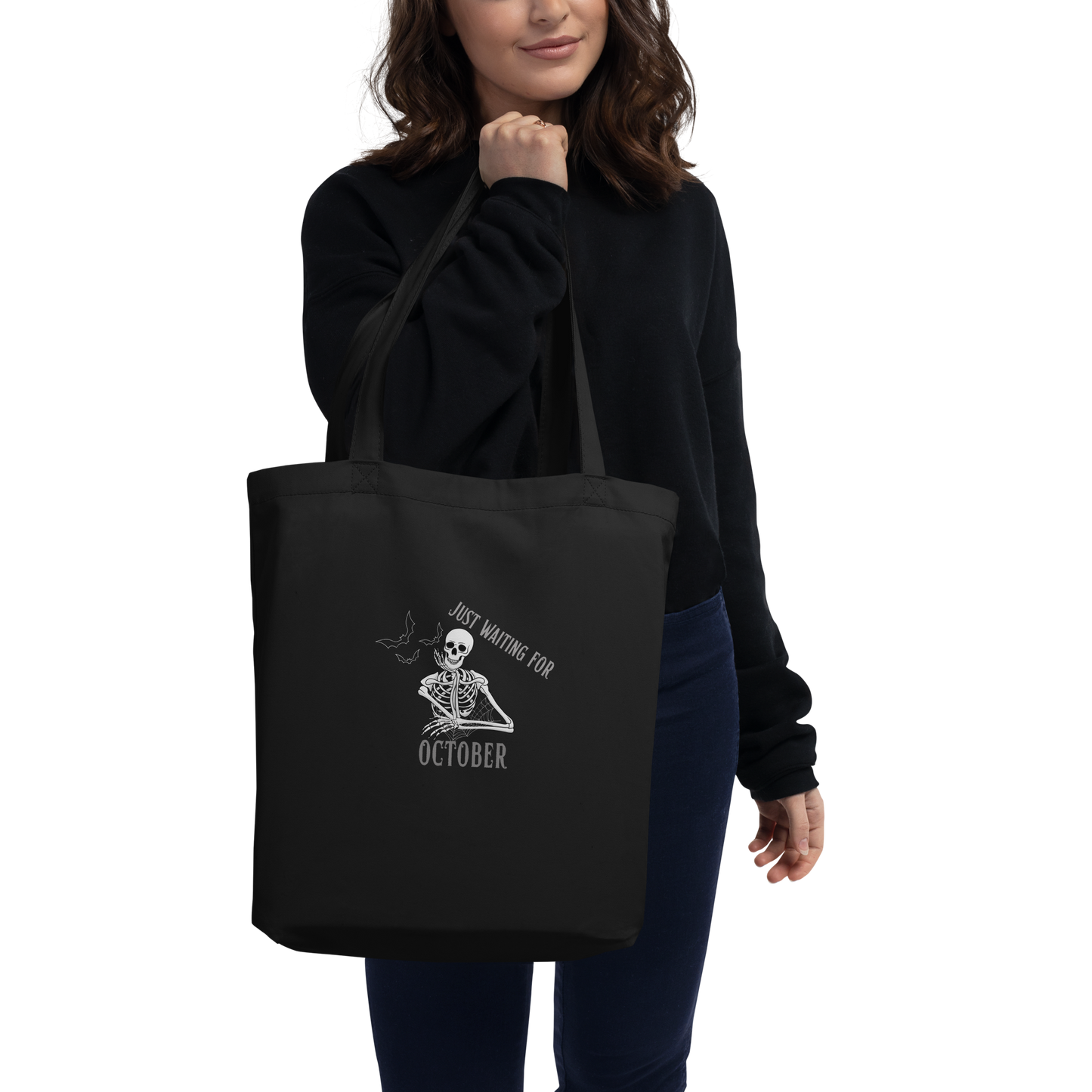 This is a woman with dark hair holding a black tote bag that has a bored skeleton with his head resting in his hand and he has spider webs and bats around him and the text reads just waiting for October
