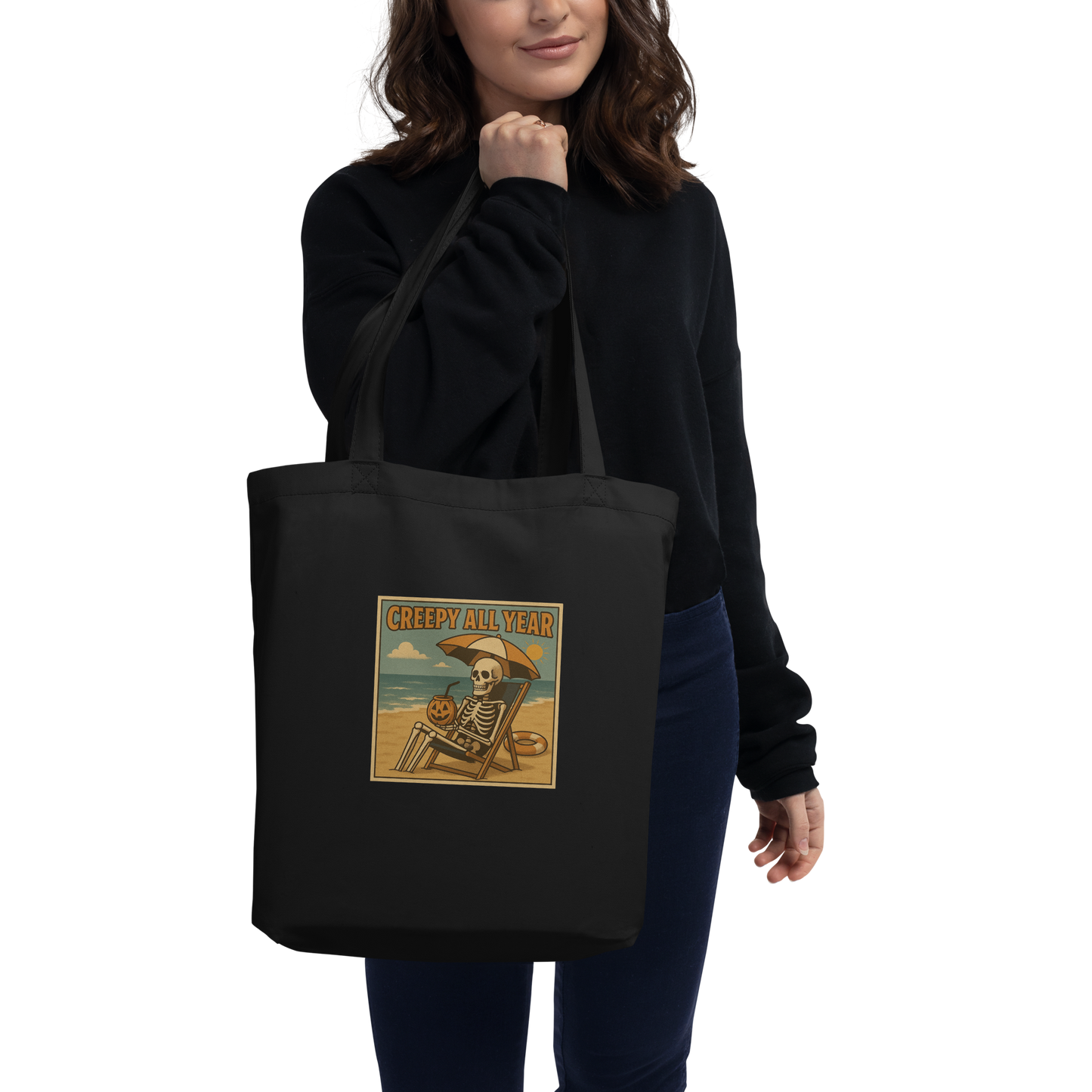 This is a woman who is holding an eco tote bag with a retro-style illustration of a grinning skeleton lounging on a beach chair under an orange umbrella. The skeleton is holding a pumpkin-shaped drink cup with a straw and relaxing by the ocean, with a bright sun in the sky and a life preserver in the sand. The words “Creepy All Year” appear boldly at the top