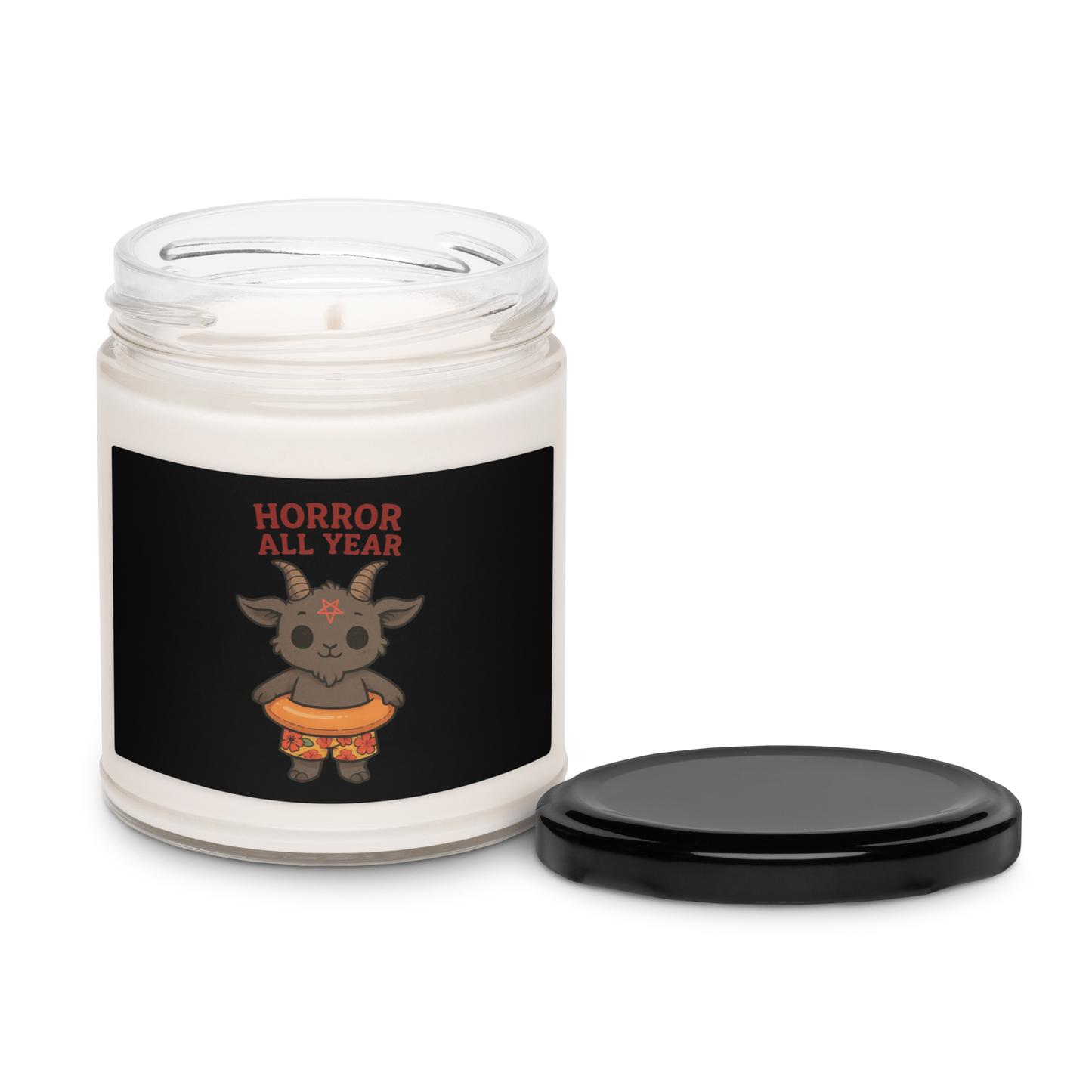 This is a white candle with a black tin lid on the front has a cartoon illustration of a baby Baphomet goat with curved horns and a playful grin, wearing bright floral swim trunks while floating in a colorful inner tube. The character is set against a light background with the bold text “Horror All Year” above in red letters