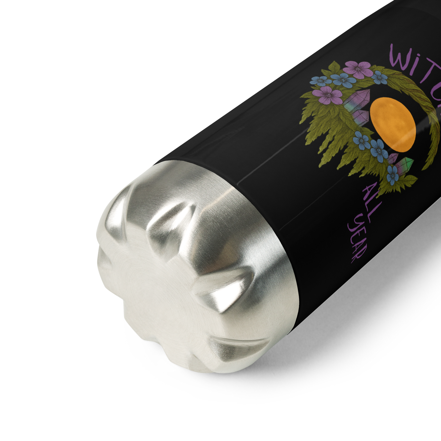 This is a zoom in of a black stainless steel water bottle with a design of a hand-drawn wreath made of moss-covered branches wraps around a glowing sun. The wreath is decorated with purple and blue flowers, small amethyst and fluorite crystals, and strands of hanging moss. The words “Witchy” curve above the wreath and “All Year” sits on the bottom right