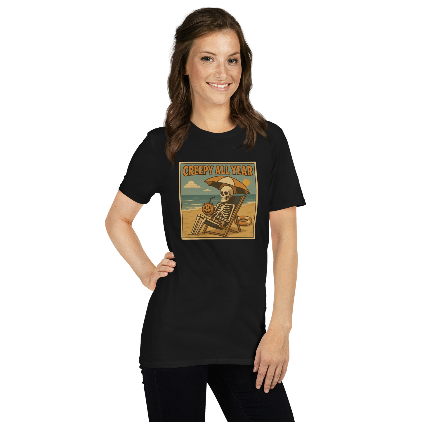 This is a woman with dark hair wearing a black t-shirt with a retro-style illustration of a grinning skeleton lounging on a beach chair under an orange umbrella. The skeleton is holding a pumpkin-shaped drink cup with a straw and relaxing by the ocean, with a bright sun in the sky and a life preserver in the sand. The words “Creepy All Year” appear boldly at the top