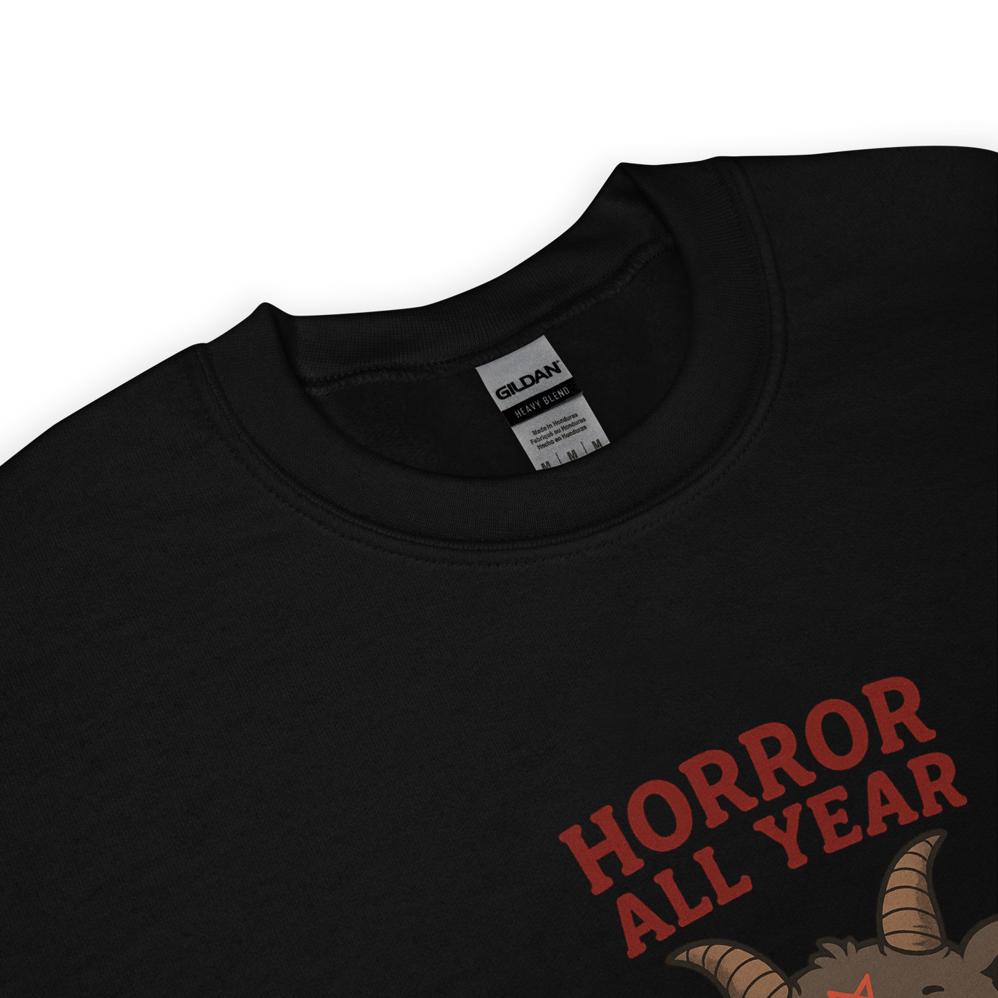 This is a zoom in of a black sweatshirt with a cartoon illustration of a baby Baphomet goat with curved horns and a playful grin, wearing bright floral swim trunks while floating in a colorful inner tube. The character is set against a light background with the bold text “Horror All Year” above in red letters