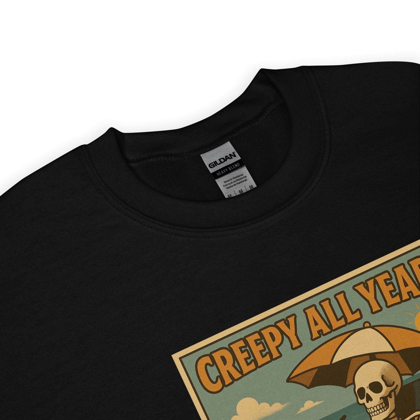 This is a zoom in of a black sweatshirt with a retro-style illustration of a grinning skeleton lounging on a beach chair under an orange umbrella. The skeleton is holding a pumpkin-shaped drink cup with a straw and relaxing by the ocean, with a bright sun in the sky and a life preserver in the sand. The words “Creepy All Year” appear boldly at the top