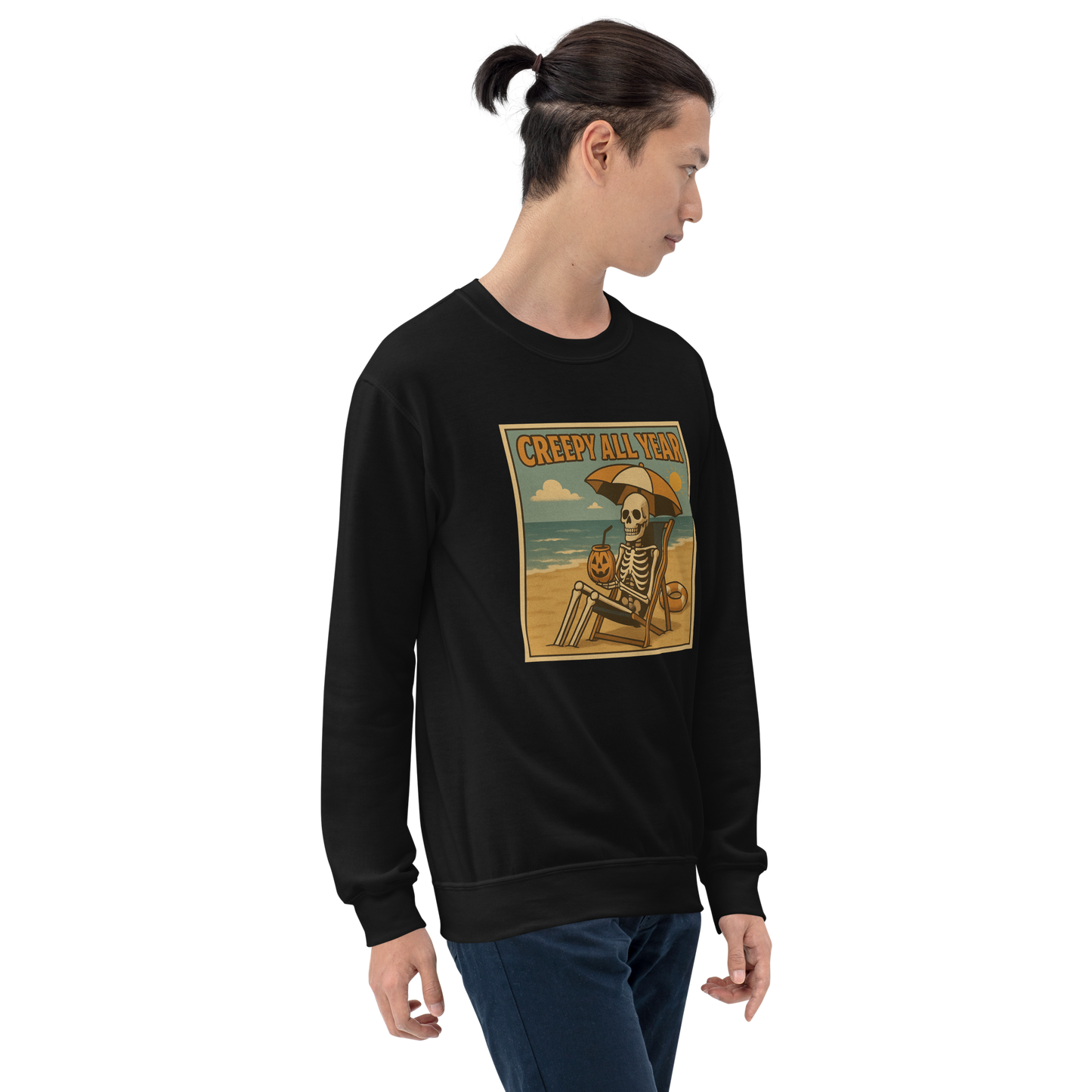 This is a man with dark hair wearing a black sweatshirt with a retro-style illustration of a grinning skeleton lounging on a beach chair under an orange umbrella. The skeleton is holding a pumpkin-shaped drink cup with a straw and relaxing by the ocean, with a bright sun in the sky and a life preserver in the sand. The words “Creepy All Year” appear boldly at the top
