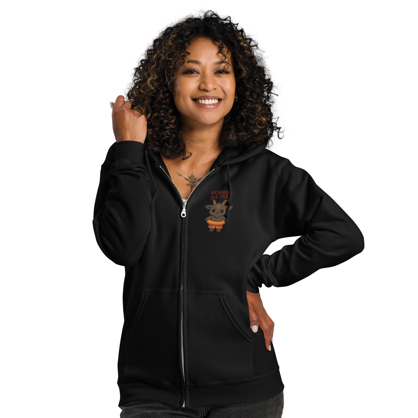 This is a woman with dark hair wearing a black zip-up hoodie with a cartoon illustration of a baby Baphomet goat with curved horns and a playful grin, wearing bright floral swim trunks while floating in a colorful inner tube. The character is set against a light background with the bold text “Horror All Year” above in red letters