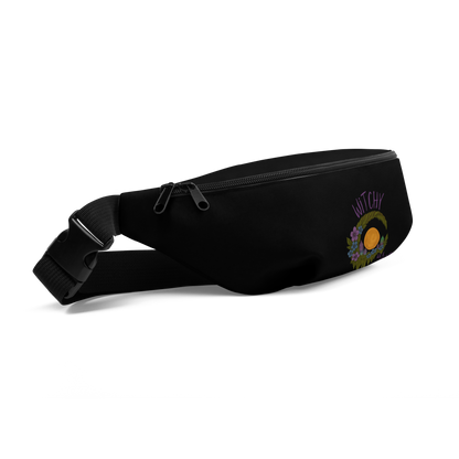 This is a black waist bag fanny pack with a design of a hand-drawn wreath made of moss-covered branches wraps around a glowing sun. The wreath is decorated with purple and blue flowers, small amethyst and fluorite crystals, and strands of hanging moss. The words “Witchy” curve above the wreath and “All Year” sits on the bottom right 