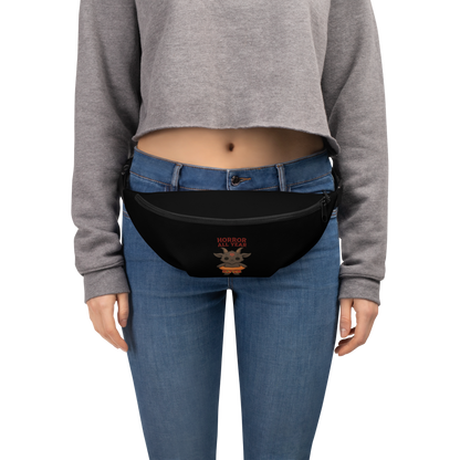 This is a woman wearing a black waist bag fanny pack that has a cartoon illustration of a baby Baphomet goat with curved horns and a playful grin, wearing bright floral swim trunks while floating in a colorful inner tube. The character is set against a light background with the bold text “Horror All Year” above in red letters 