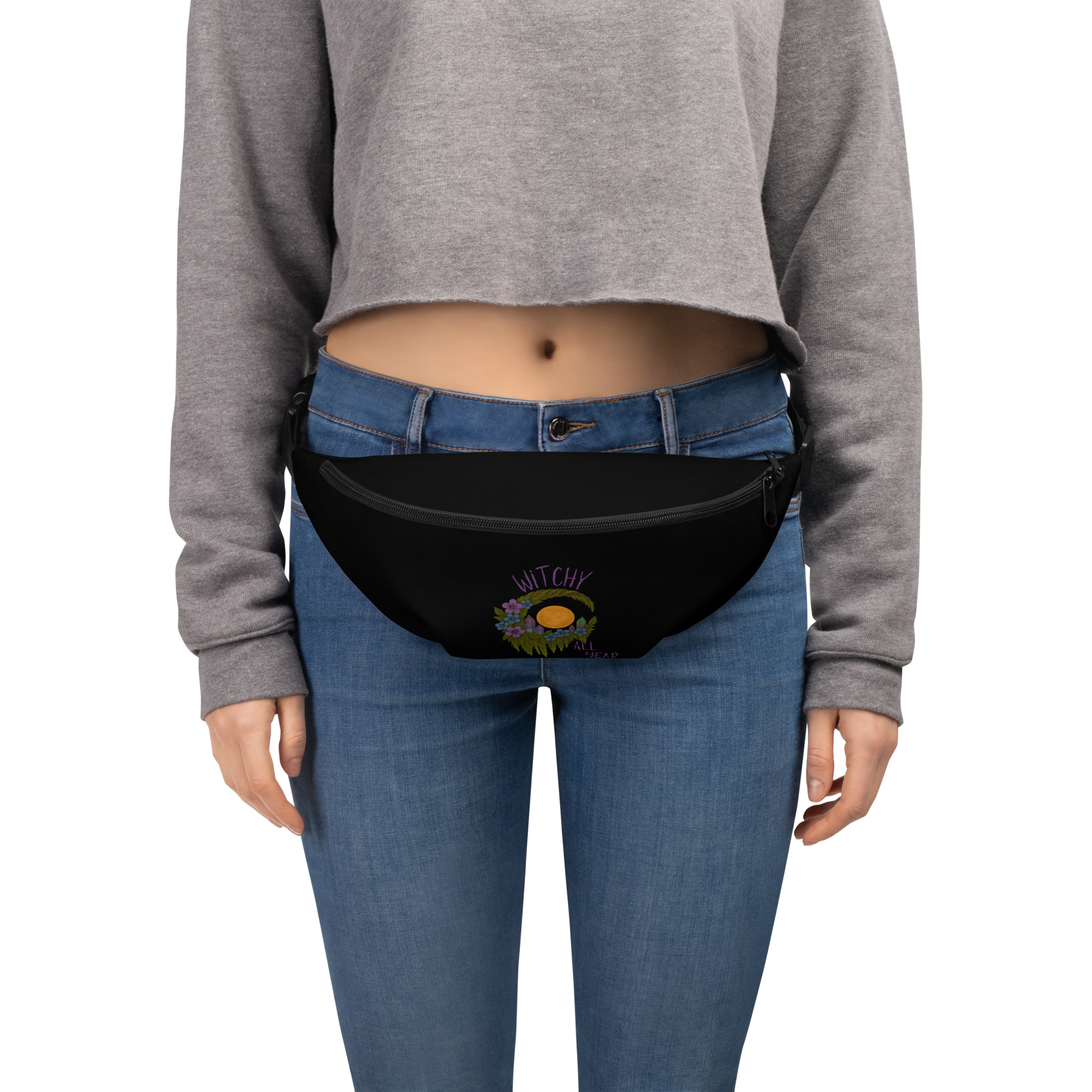 This is a woman wearing a black waist bag fanny pack with a design of a hand-drawn wreath made of moss-covered branches wraps around a glowing sun. The wreath is decorated with purple and blue flowers, small amethyst and fluorite crystals, and strands of hanging moss. The words “Witchy” curve above the wreath and “All Year” sits on the bottom right 
