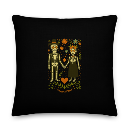 This is a black throw pillow with a Día de los Muertos–style illustration of a male and female skeleton holding hands, surrounded by marigolds, vines, and folk art patterns. The skeletons wear traditional black clothing, the man in a suit and hat, the woman with a floral crown with a red heart and leaves at the bottom and the text Creepy All Year. Rendered in warm tones of orange, green, and cream, symbolizing eternal love and remembrance 