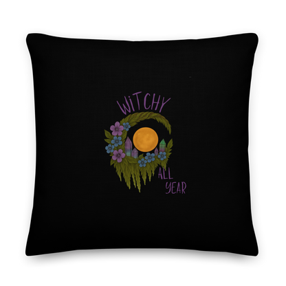 This is a black pillow with a design of a hand-drawn wreath made of moss-covered branches wraps around a glowing sun. The wreath is decorated with purple and blue flowers, small amethyst and fluorite crystals, and strands of hanging moss. The words “Witchy” curve above the wreath and “All Year” sits on the bottom right 