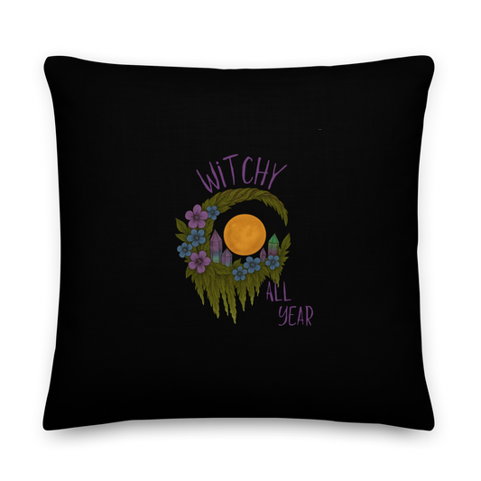 This is a black pillow with a design of a hand-drawn wreath made of moss-covered branches wraps around a glowing sun. The wreath is decorated with purple and blue flowers, small amethyst and fluorite crystals, and strands of hanging moss. The words “Witchy” curve above the wreath and “All Year” sits on the bottom right 