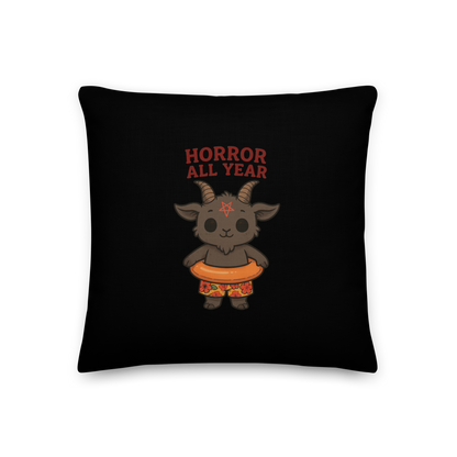 This is a black pillow and on the front has a cartoon illustration of a baby Baphomet goat with curved horns and a playful grin, wearing bright floral swim trunks while floating in a colorful inner tube. The character is set against a light background with the bold text “Horror All Year” above in red letters