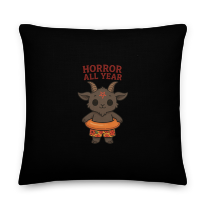 This is a black throw pillow and on the front has a cartoon illustration of a baby Baphomet goat with curved horns and a playful grin, wearing bright floral swim trunks while floating in a colorful inner tube. The character is set against a light background with the bold text “Horror All Year” above in red letters