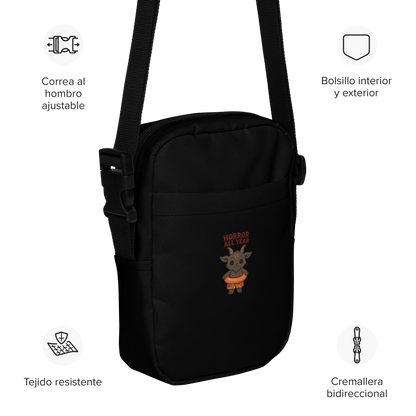 This is a black crossbody bag with a cartoon illustration of a baby Baphomet goat with curved horns and a playful grin, wearing bright floral swim trunks while floating in a colorful inner tube. The character is set against a light background with the bold text “Horror All Year” above in red letters