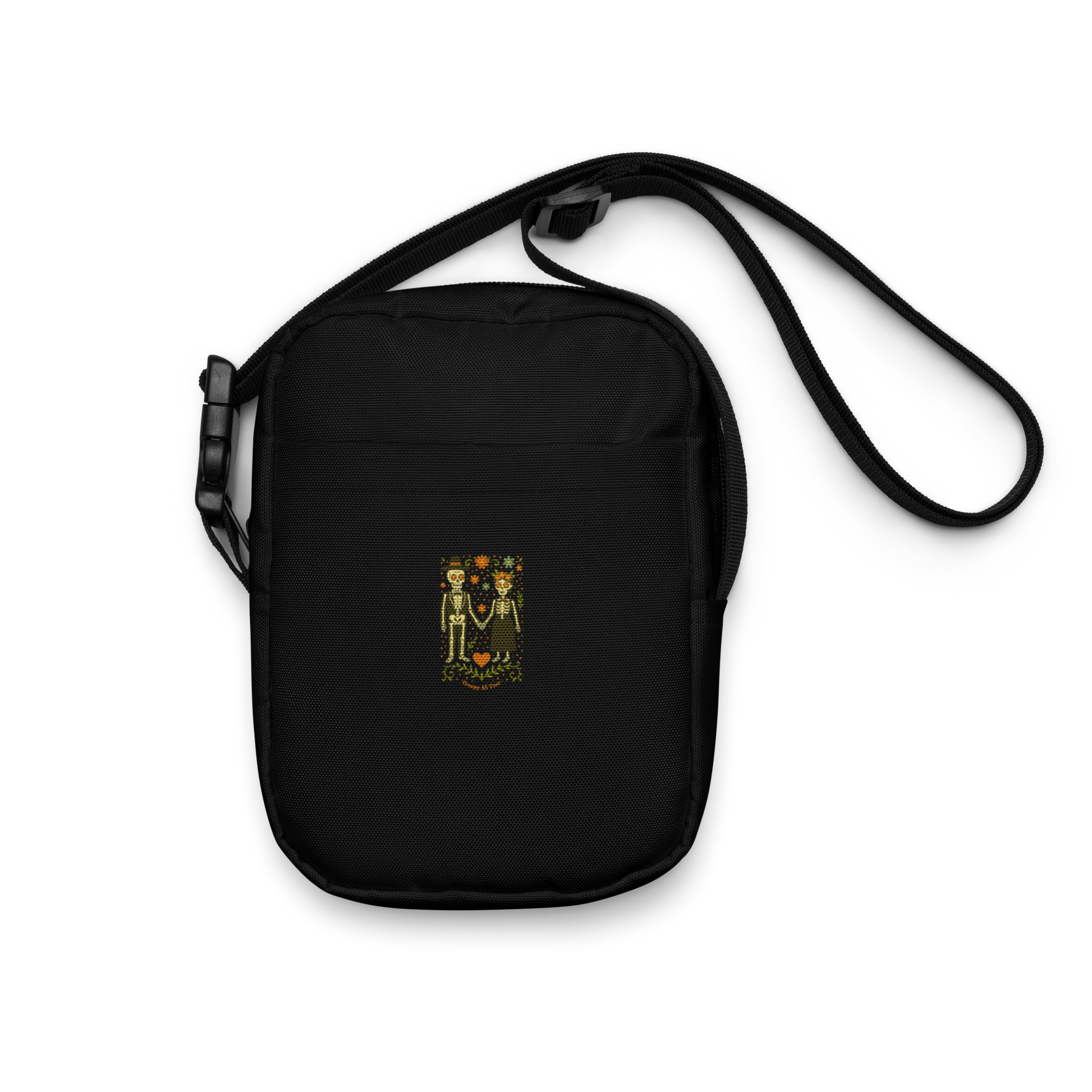 This is a black crossbody bag with a Día de los Muertos–style illustration of a male and female skeleton holding hands, surrounded by marigolds, vines, and folk art patterns. The skeletons wear traditional black clothing, the man in a suit and hat, the woman with a floral crown with a red heart and leaves at the bottom and the text Creepy All Year. Rendered in warm tones of orange, green, and cream, symbolizing eternal love and remembrance
