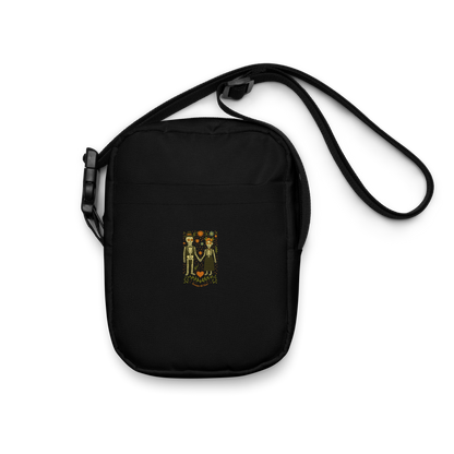 This is a black crossbody bag with a Día de los Muertos–style illustration of a male and female skeleton holding hands, surrounded by marigolds, vines, and folk art patterns. The skeletons wear traditional black clothing, the man in a suit and hat, the woman with a floral crown with a red heart and leaves at the bottom and the text Creepy All Year. Rendered in warm tones of orange, green, and cream, symbolizing eternal love and remembrance