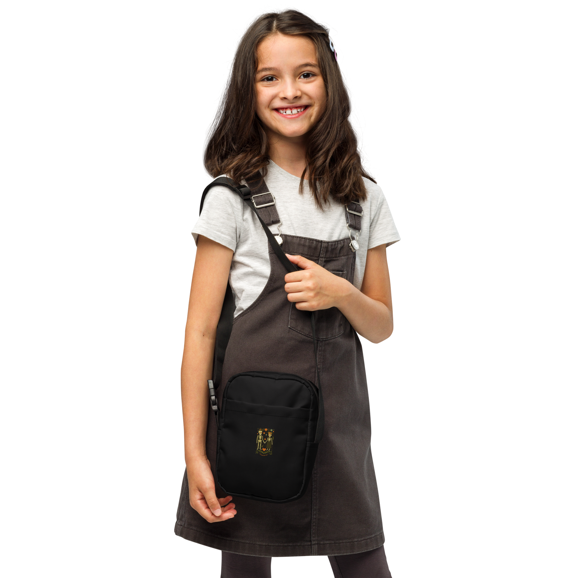 This is a girl with dark hair wearing a black crossbody bag with a Día de los Muertos–style illustration of a male and female skeleton holding hands, surrounded by marigolds, vines, and folk art patterns. The skeletons wear traditional black clothing, the man in a suit and hat, the woman with a floral crown with a red heart and leaves at the bottom and the text Creepy All Year. Rendered in warm tones of orange, green, and cream, symbolizing eternal love and remembrance