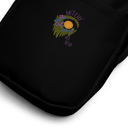 This is a zoom in of a black crossbody bag with a design of a hand-drawn wreath made of moss-covered branches wraps around a glowing sun. The wreath is decorated with purple and blue flowers, small amethyst and fluorite crystals, and strands of hanging moss. The words “Witchy” curve above the wreath and “All Year” sits on the bottom right 