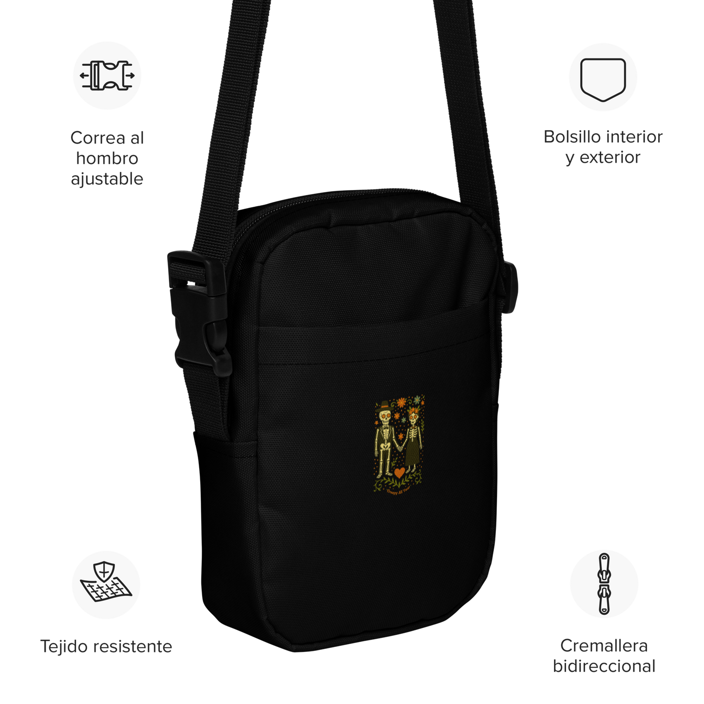This is a black crossbody bag with a Día de los Muertos–style illustration of a male and female skeleton holding hands, surrounded by marigolds, vines, and folk art patterns. The skeletons wear traditional black clothing, the man in a suit and hat, the woman with a floral crown with a red heart and leaves at the bottom and the text Creepy All Year. Rendered in warm tones of orange, green, and cream, symbolizing eternal love and remembrance