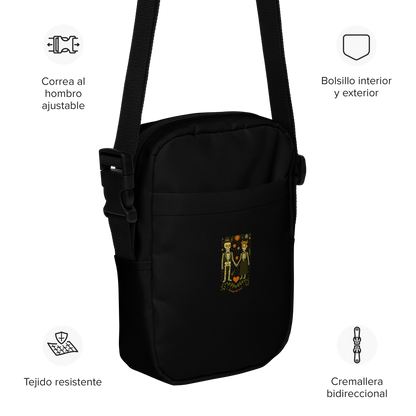 This is a black crossbody bag with a Día de los Muertos–style illustration of a male and female skeleton holding hands, surrounded by marigolds, vines, and folk art patterns. The skeletons wear traditional black clothing, the man in a suit and hat, the woman with a floral crown with a red heart and leaves at the bottom and the text Creepy All Year. Rendered in warm tones of orange, green, and cream, symbolizing eternal love and remembrance