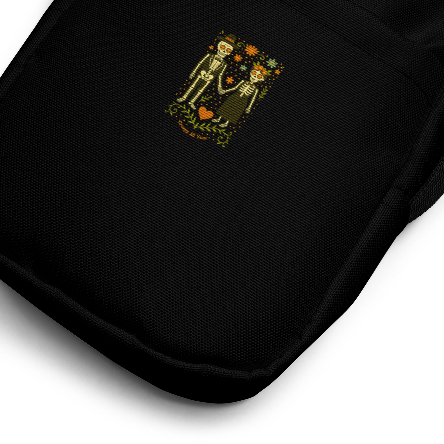 This is a zoom in of a black crossbody bag with a Día de los Muertos–style illustration of a male and female skeleton holding hands, surrounded by marigolds, vines, and folk art patterns. The skeletons wear traditional black clothing, the man in a suit and hat, the woman with a floral crown with a red heart and leaves at the bottom and the text Creepy All Year. Rendered in warm tones of orange, green, and cream, symbolizing eternal love and remembrance