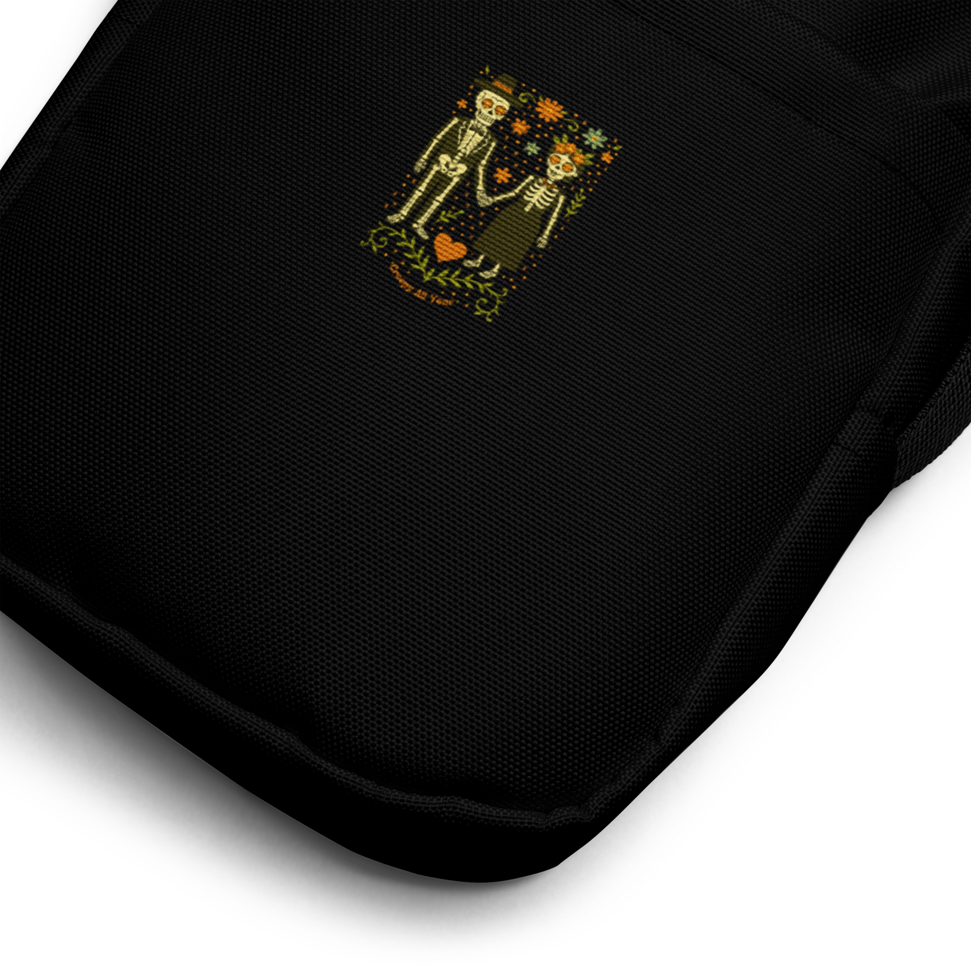 This is a zoom in of a black crossbody bag with a Día de los Muertos–style illustration of a male and female skeleton holding hands, surrounded by marigolds, vines, and folk art patterns. The skeletons wear traditional black clothing, the man in a suit and hat, the woman with a floral crown with a red heart and leaves at the bottom and the text Creepy All Year. Rendered in warm tones of orange, green, and cream, symbolizing eternal love and remembrance