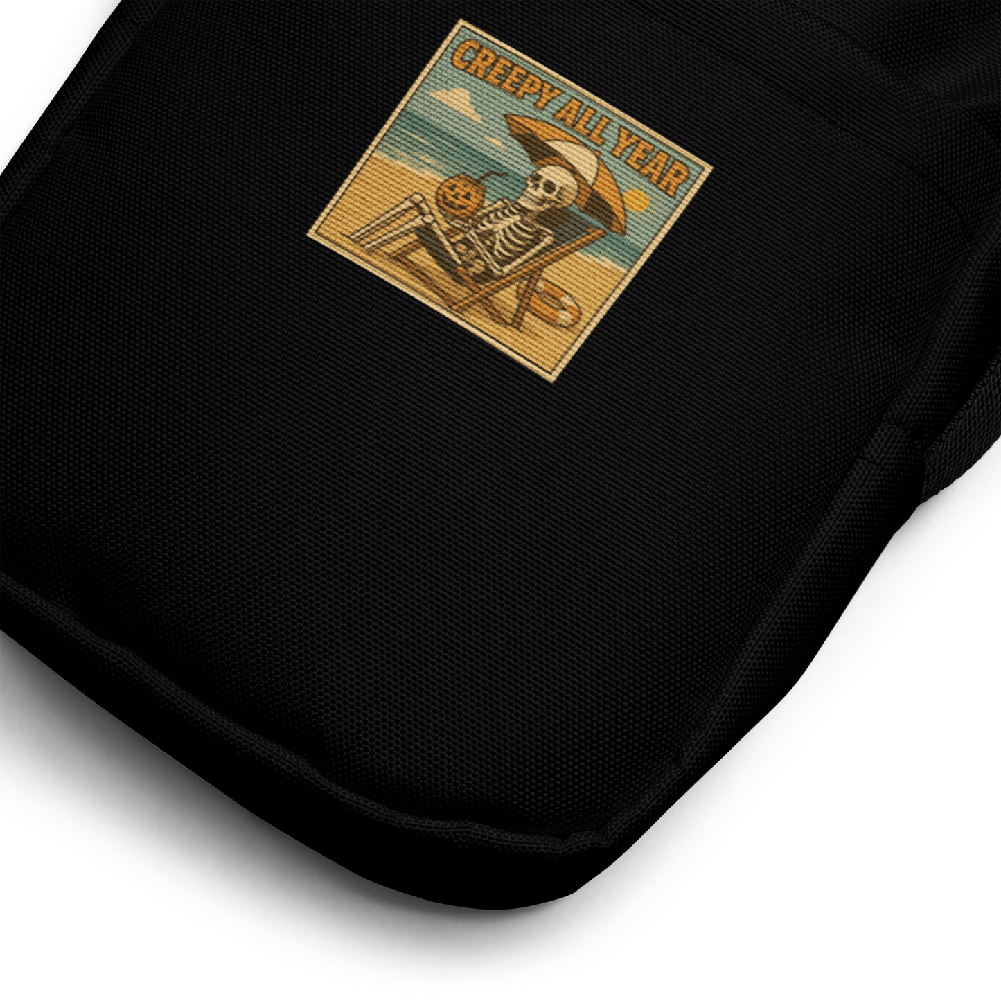 This is a zoom in of a black crossbody bag with a retro-style illustration of a grinning skeleton lounging on a beach chair under an orange umbrella. The skeleton is holding a pumpkin-shaped drink cup with a straw and relaxing by the ocean, with a bright sun in the sky and a life preserver in the sand. The words “Creepy All Year” appear boldly at the top 