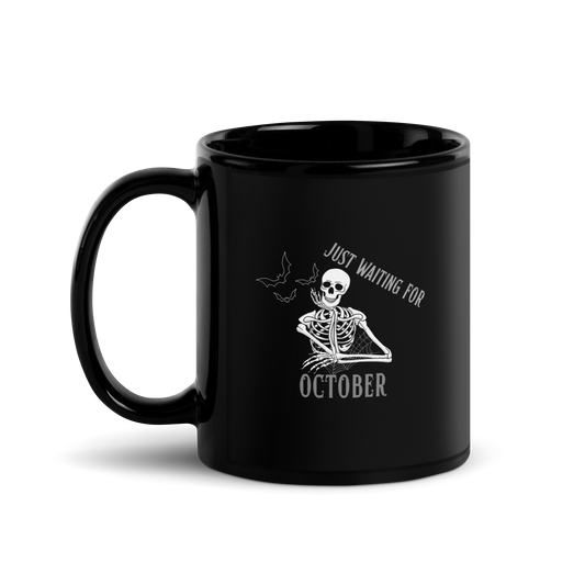 This is a black mug that has a bored skeleton with his head resting in his hand and he has spider webs and bats around him and the text reads just waiting for October