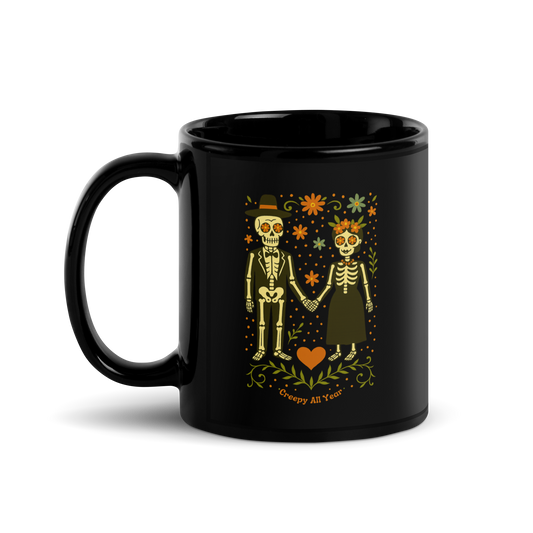 This is a black mug with a Día de los Muertos–style illustration of a male and female skeleton holding hands, surrounded by marigolds, vines, and folk art patterns. The skeletons wear traditional black clothing, the man in a suit and hat, the woman with a floral crown with a red heart and leaves at the bottom and the text Creepy All Year. Rendered in warm tones of orange, green, and cream, symbolizing eternal love and remembrance