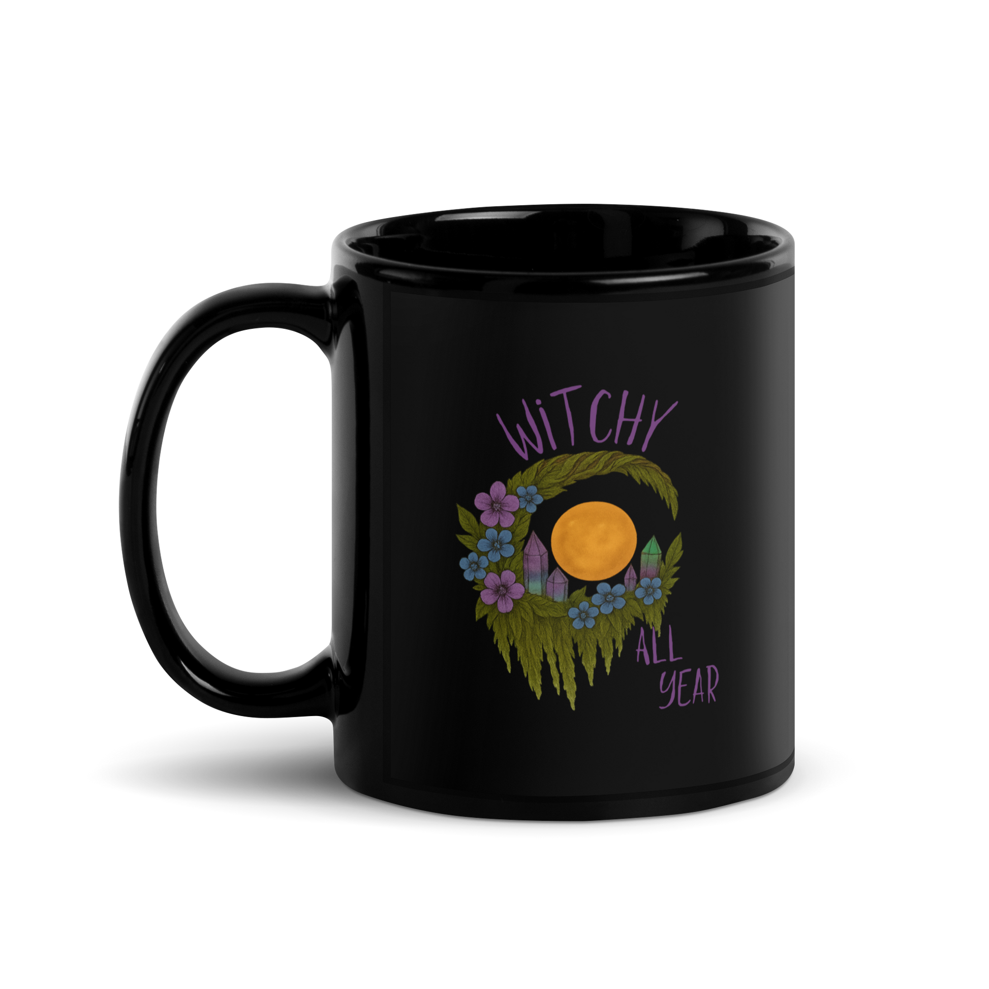 This is a black glossy mug with a design of a hand-drawn wreath made of moss-covered branches wraps around a glowing sun. The wreath is decorated with purple and blue flowers, small amethyst and fluorite crystals, and strands of hanging moss. The words “Witchy” curve above the wreath and “All Year” sits on the bottom right  