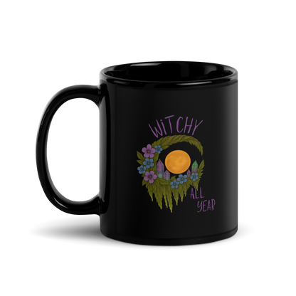 This is a black glossy mug with a design of a hand-drawn wreath made of moss-covered branches wraps around a glowing sun. The wreath is decorated with purple and blue flowers, small amethyst and fluorite crystals, and strands of hanging moss. The words “Witchy” curve above the wreath and “All Year” sits on the bottom right  