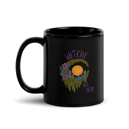 This is a black glossy mug with a design of a hand-drawn wreath made of moss-covered branches wraps around a glowing sun. The wreath is decorated with purple and blue flowers, small amethyst and fluorite crystals, and strands of hanging moss. The words “Witchy” curve above the wreath and “All Year” sits on the bottom right  