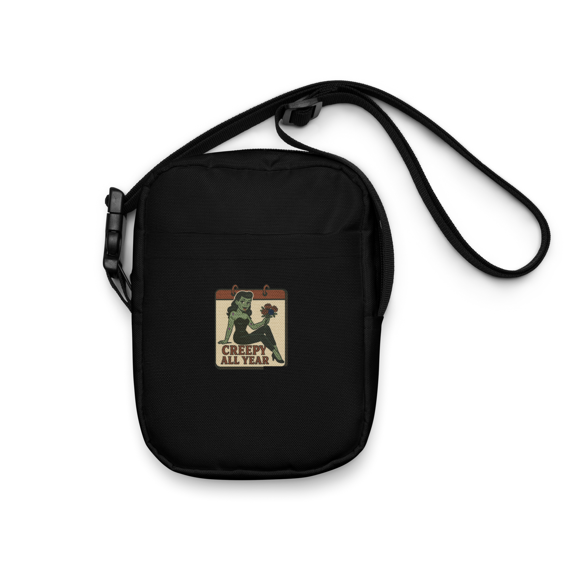 Black crossbody bag with a graphic design. The design features a green pinup-style zombie girl holding flowers, with a banner underneath that reads “CREEPY ALL YEAR.”
