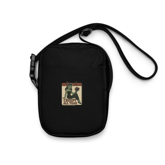 Black crossbody bag with a graphic design. The design features a green pinup-style zombie girl holding flowers, with a banner underneath that reads “CREEPY ALL YEAR.”