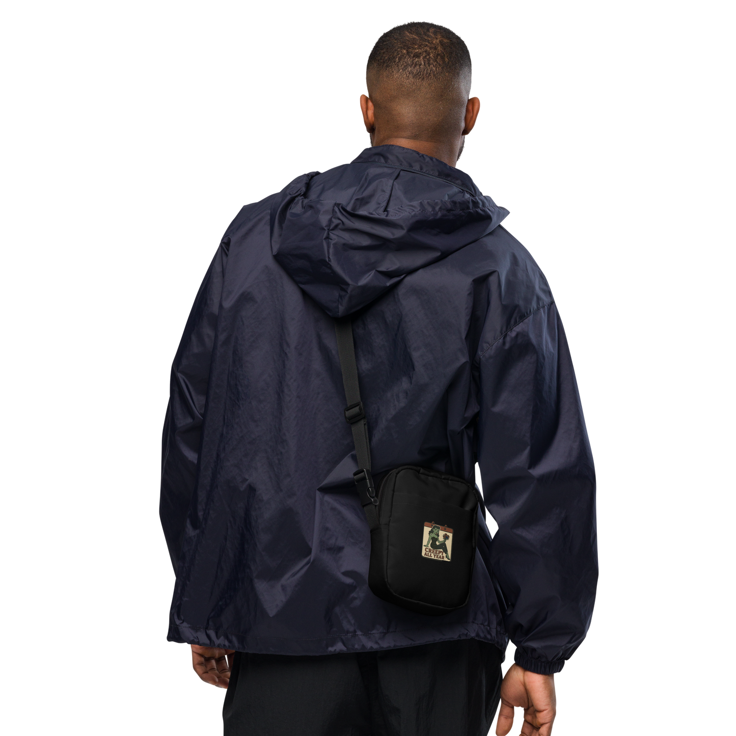 Person wearing a navy raincoat with a black crossbody bag on a white background. The design features a green pinup-style zombie girl holding flowers, with a banner underneath that reads “CREEPY ALL YEAR.”