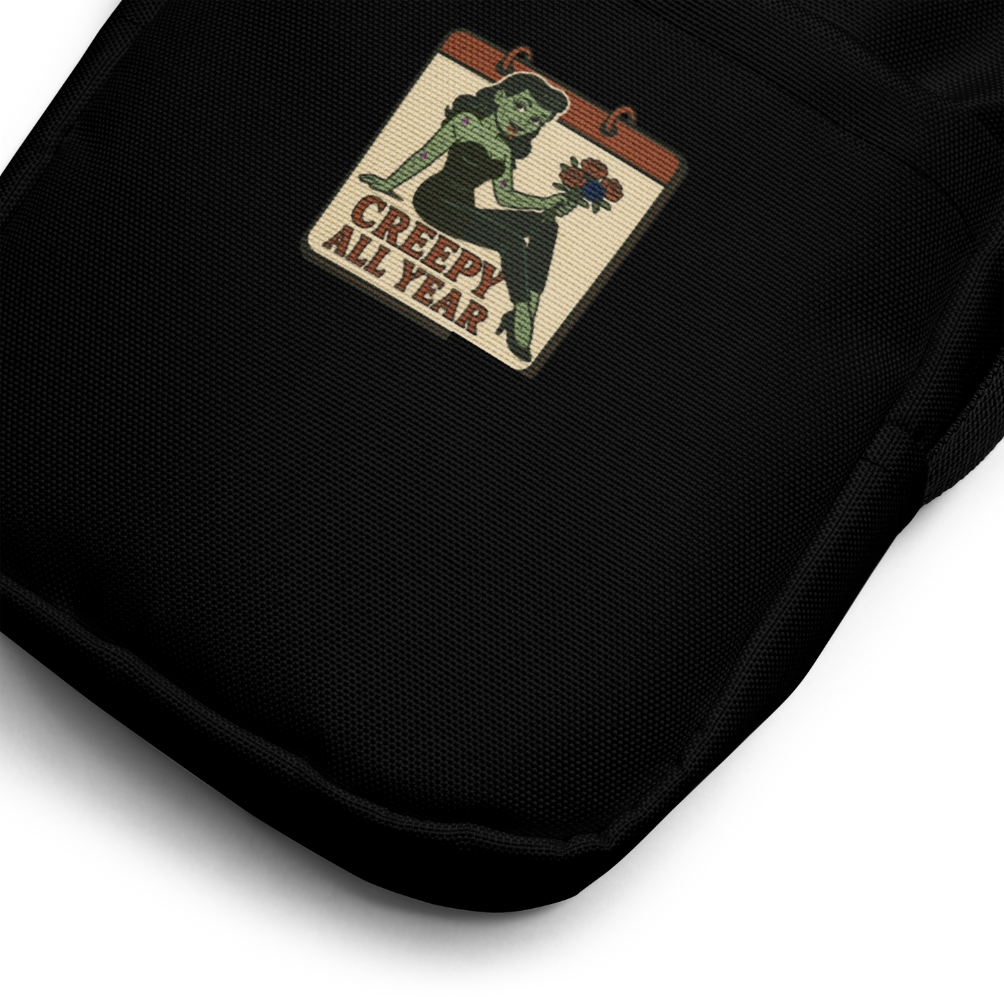 Black crossbody bag with a graphic design zoomed in. The design features a green pinup-style zombie girl holding flowers, with a banner underneath that reads “CREEPY ALL YEAR.”